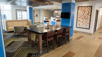 A modern hotel lobby with a reception desk, a seating area with a sofa and chairs, a dining table with chairs, and a colorful abstract painting on the wall.
