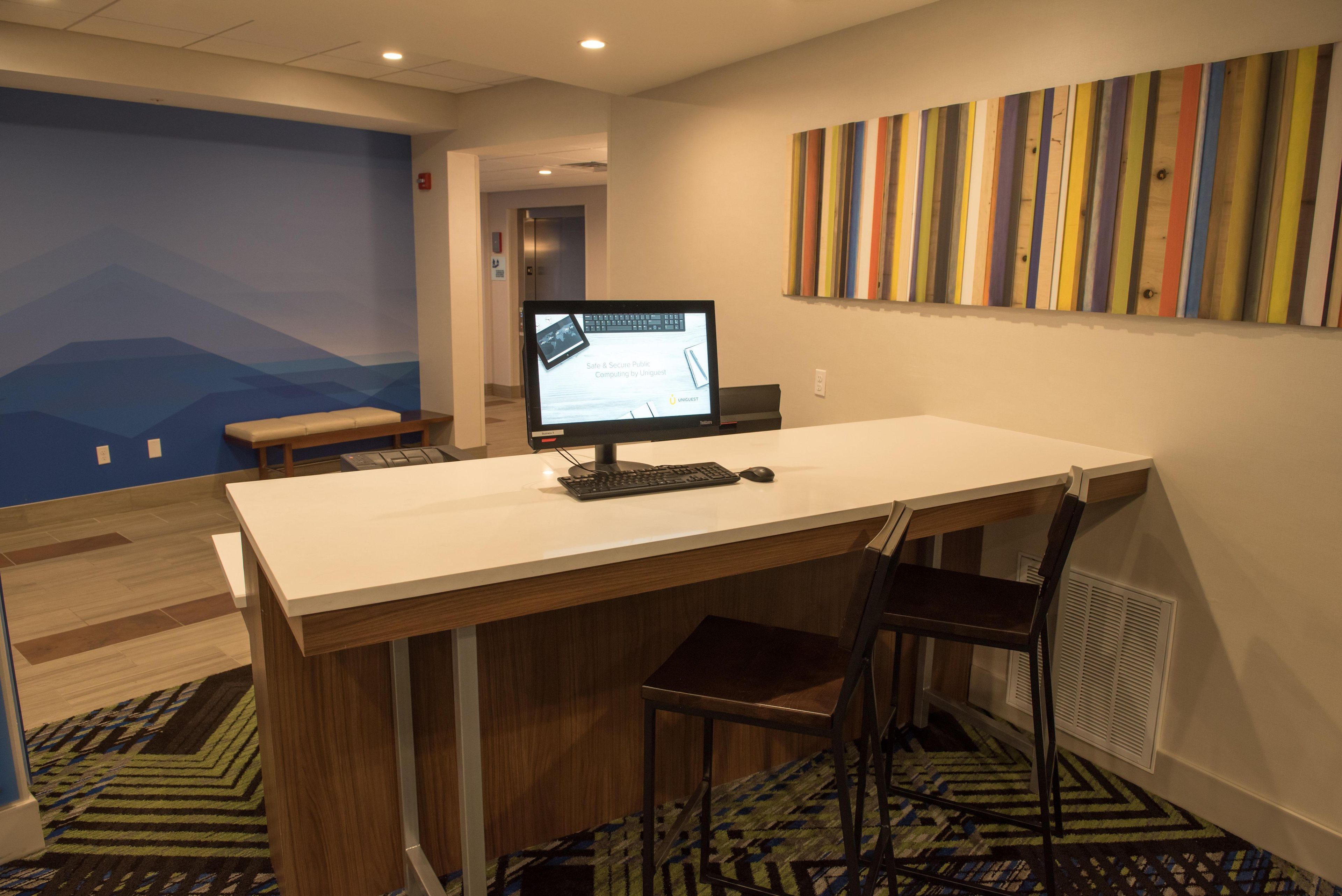 A modern office space with a desk, two chairs, a computer monitor, and a colorful striped wall art piece.
