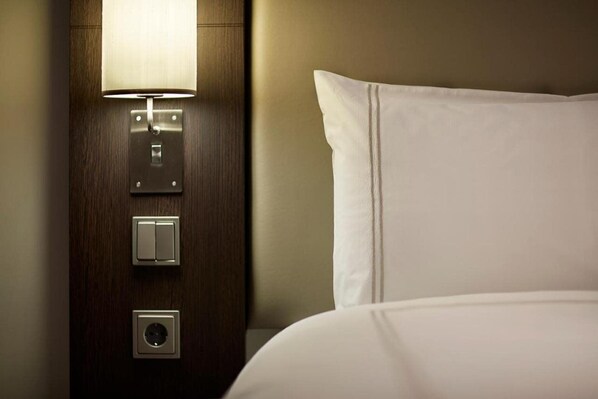 A hotel room with a wooden headboard, a bedside lamp, a power outlet, and a white pillow. - Premier Inn Munich Airport Sud Hotel (Hallbergmoos)