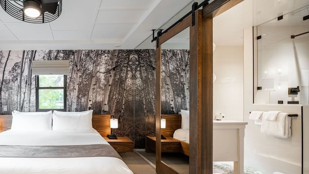 A modern hotel room with a large bed, a wooden headboard, a nightstand with a lamp, a mirror, and a bathroom with a shower and a towel rack.