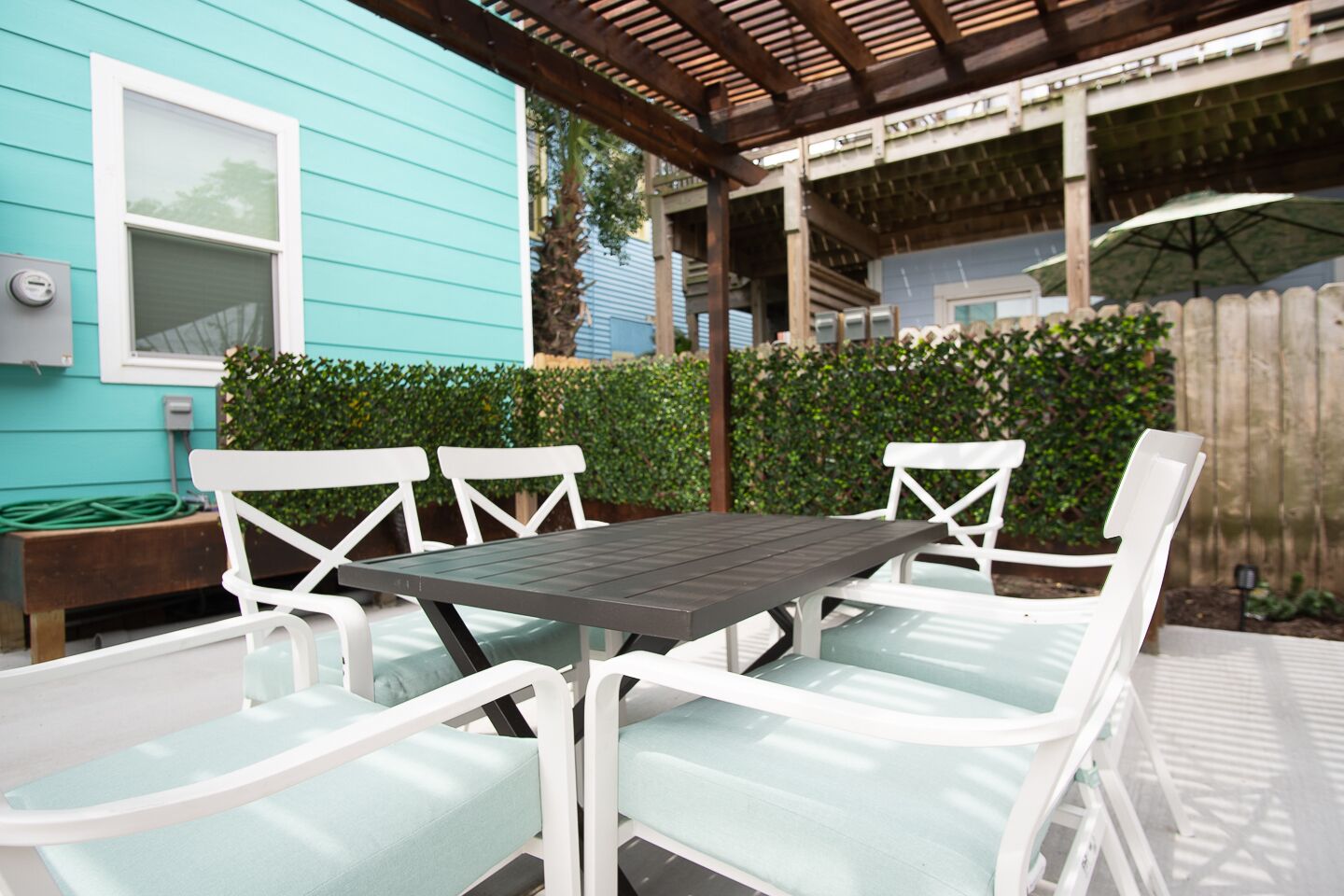 A patio with a table and white chairs, a blue house in the background, and a wooden fence.