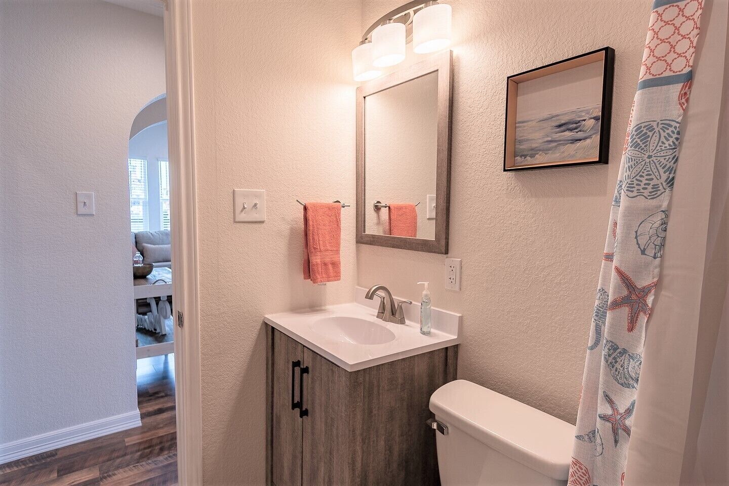 A bathroom with a white sink, wooden cabinet, and a toilet. There is a towel hanging on the wall and a framed picture above the mirror.
