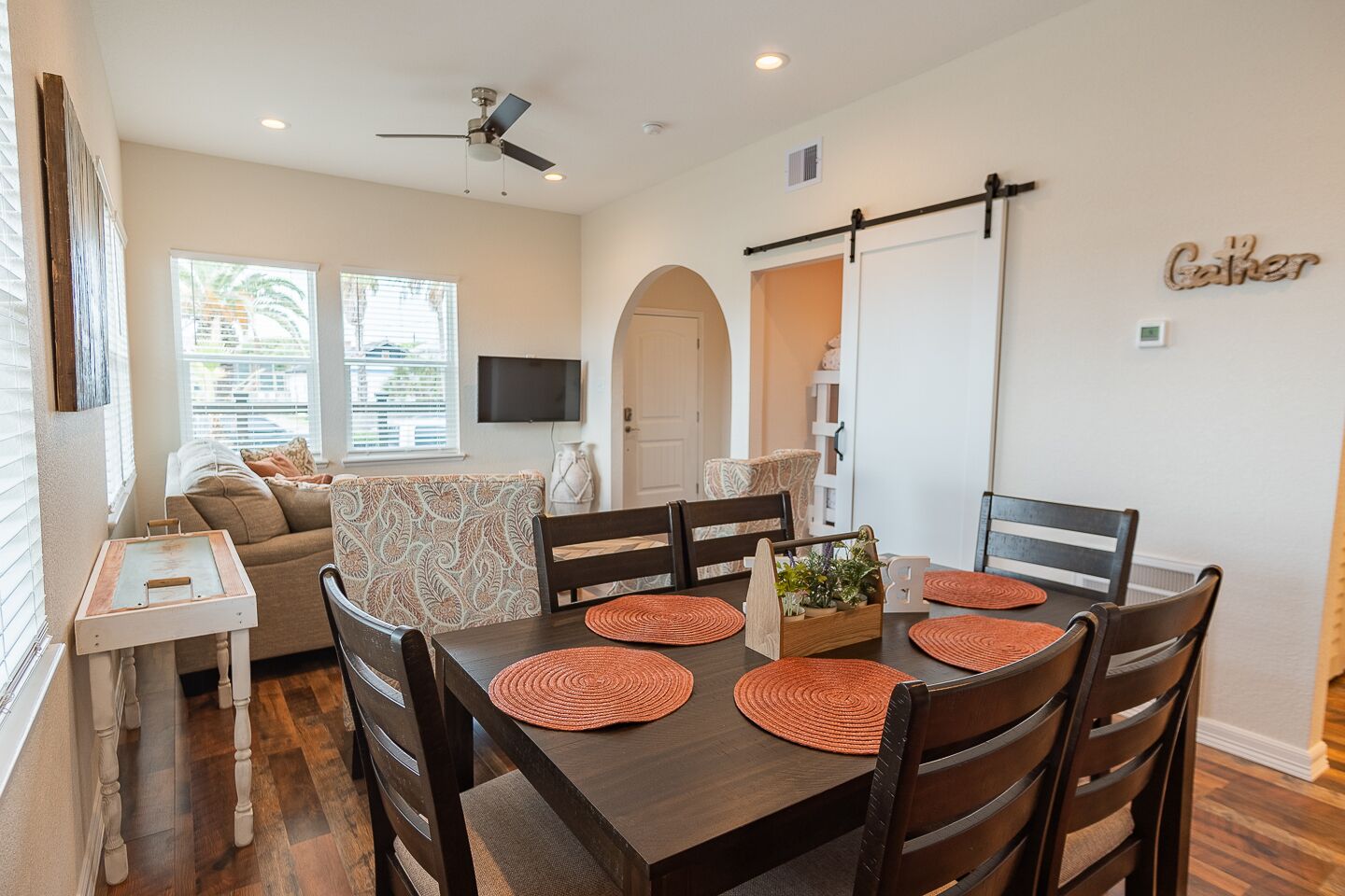A dining area with a dark wooden table, six chairs, and orange placemats. There is a television on the wall, a sofa with patterned cushions, and a "Gather" sign on the wall.