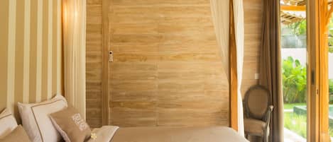 A bedroom with a wooden door, a bed with beige bedding, and a view of greenery outside.