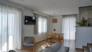 A modern living room with a sofa, a dining table with chairs, a TV mounted on the wall, and a staircase leading to the upper floor.