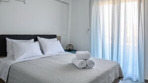 A neatly made bed with white linens and rolled towels, a dark headboard, and a window with sheer curtains.