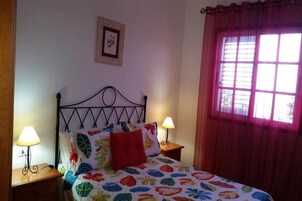 Una cama con una colcha floral, dos lámparas de noche, un cuadro en la pared y una ventana con cortina roja.