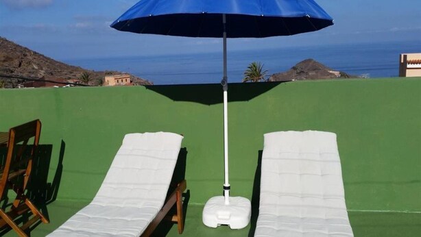Una tumbona bajo un parasol azul sobre una superficie verde, con vista al mar y colinas.