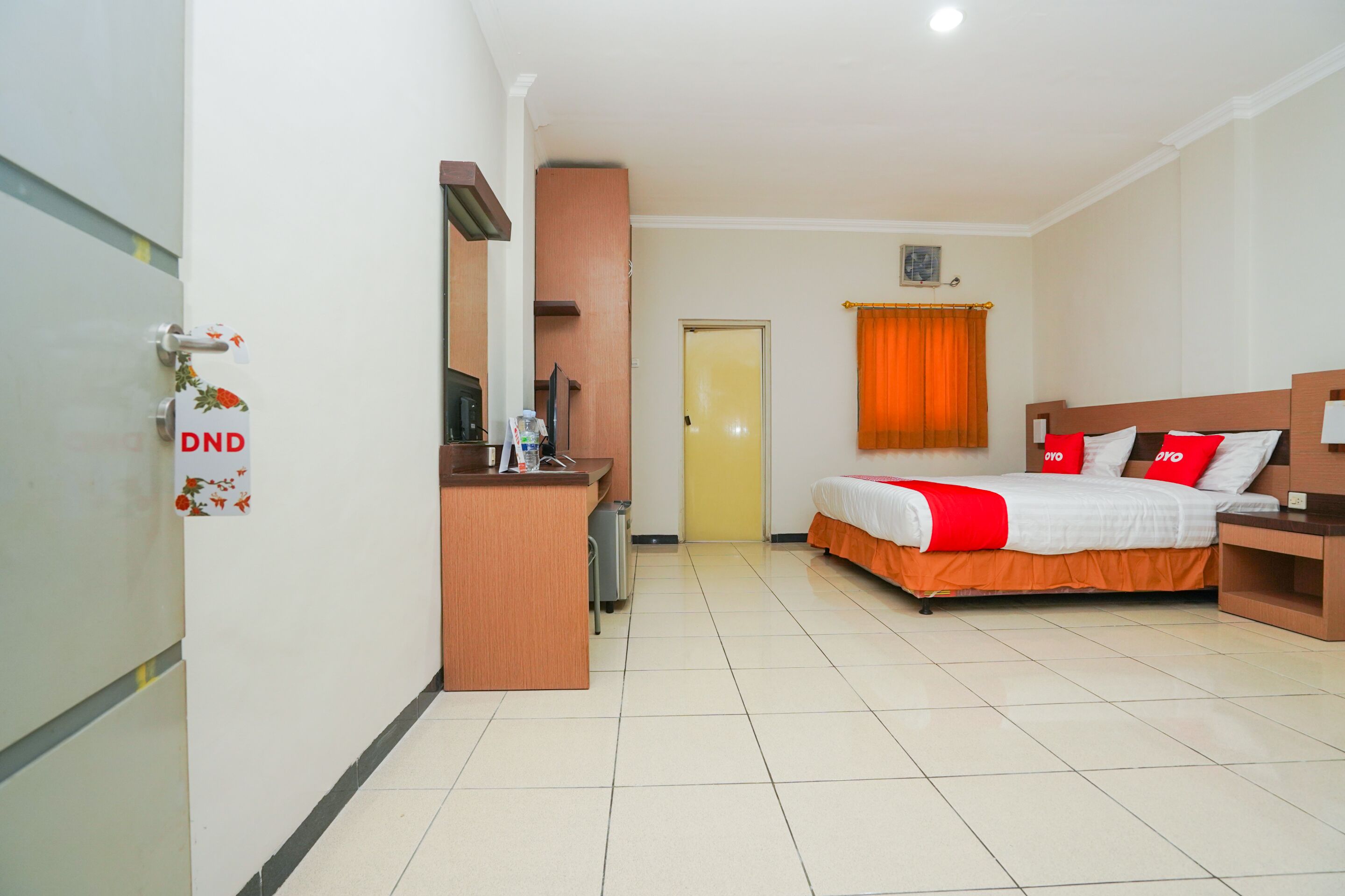 A hotel room with a bed, a desk, a chair, a wardrobe, and a door with a sign.