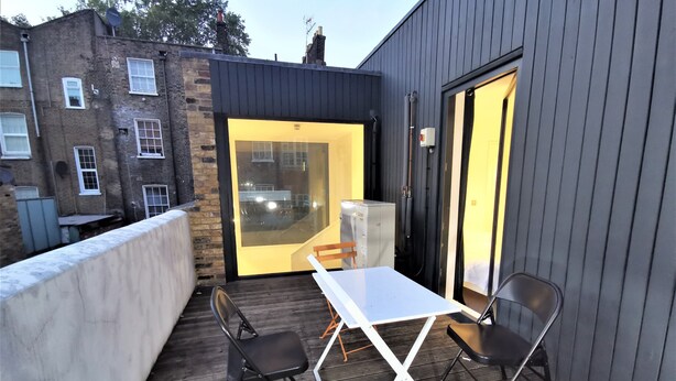 A balcony with a white table and two chairs, a glass door leading to a room, and a brick building in the background. - Double Room with Balcony - 3c (London)