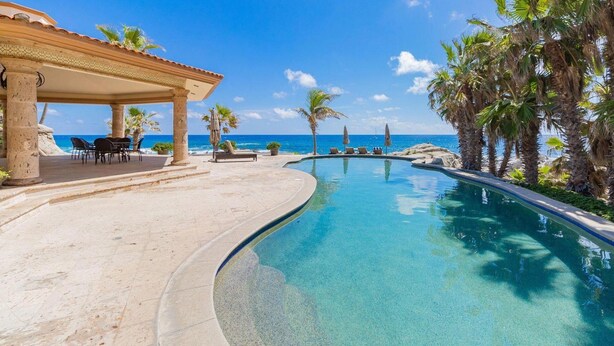 A poolside area with a curved pool, lounge chairs, and a view of the ocean. - Private Luxury Escape, Cabo San Lucas Villa 1048 (Cabo San Lucas)