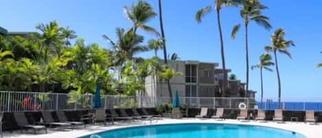 A swimming pool surrounded by lounge chairs and palm trees, with a building in the background.