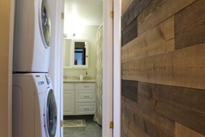 Ein Waschbereich mit Waschmaschine und Trockner, ein Badezimmer mit Waschbecken und Spiegel sowie eine Holzwand.