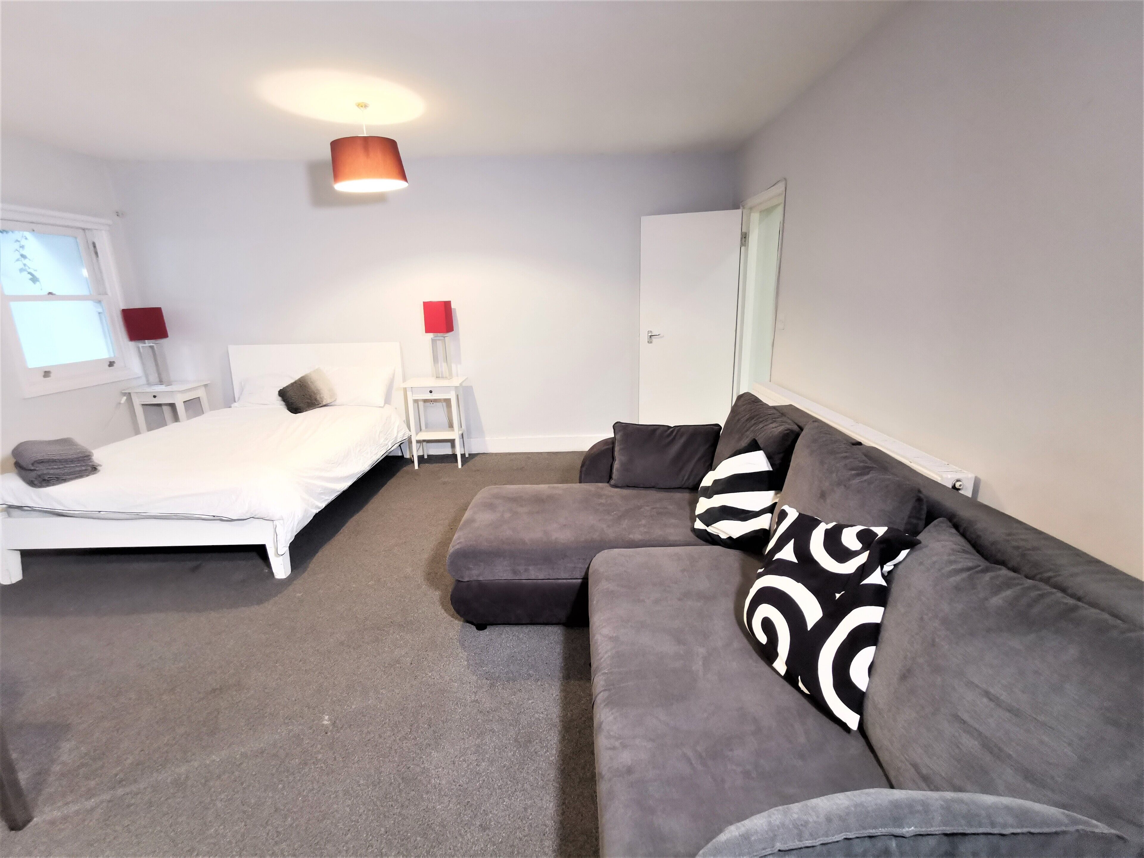 Ein Schlafzimmer mit einem Bett, einem grauen Sofa, einem kleinen Tisch, einer roten Lampe und einem weißen Kleiderschrank.
