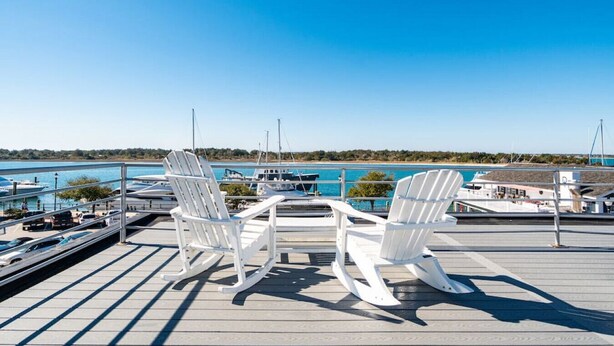 Una terraza con dos sillas Adirondack blancas y una mesa, con vista a un puerto deportivo con barcos y un cielo azul despejado.