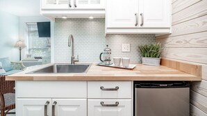 Una cocina con armarios blancos, un lavamanos de acero inoxidable y un lavavajillas. Hay una ventana con persianas, una planta y una cafetera en la encimera.