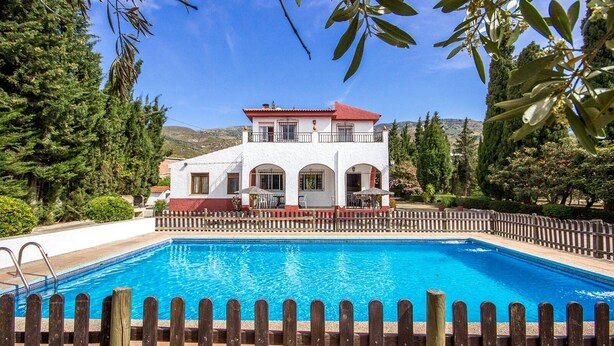 A swimming pool with a wooden deck and a white building with red accents in the background. - Sortes Ana Órgiva by Ruralidays (Granada)