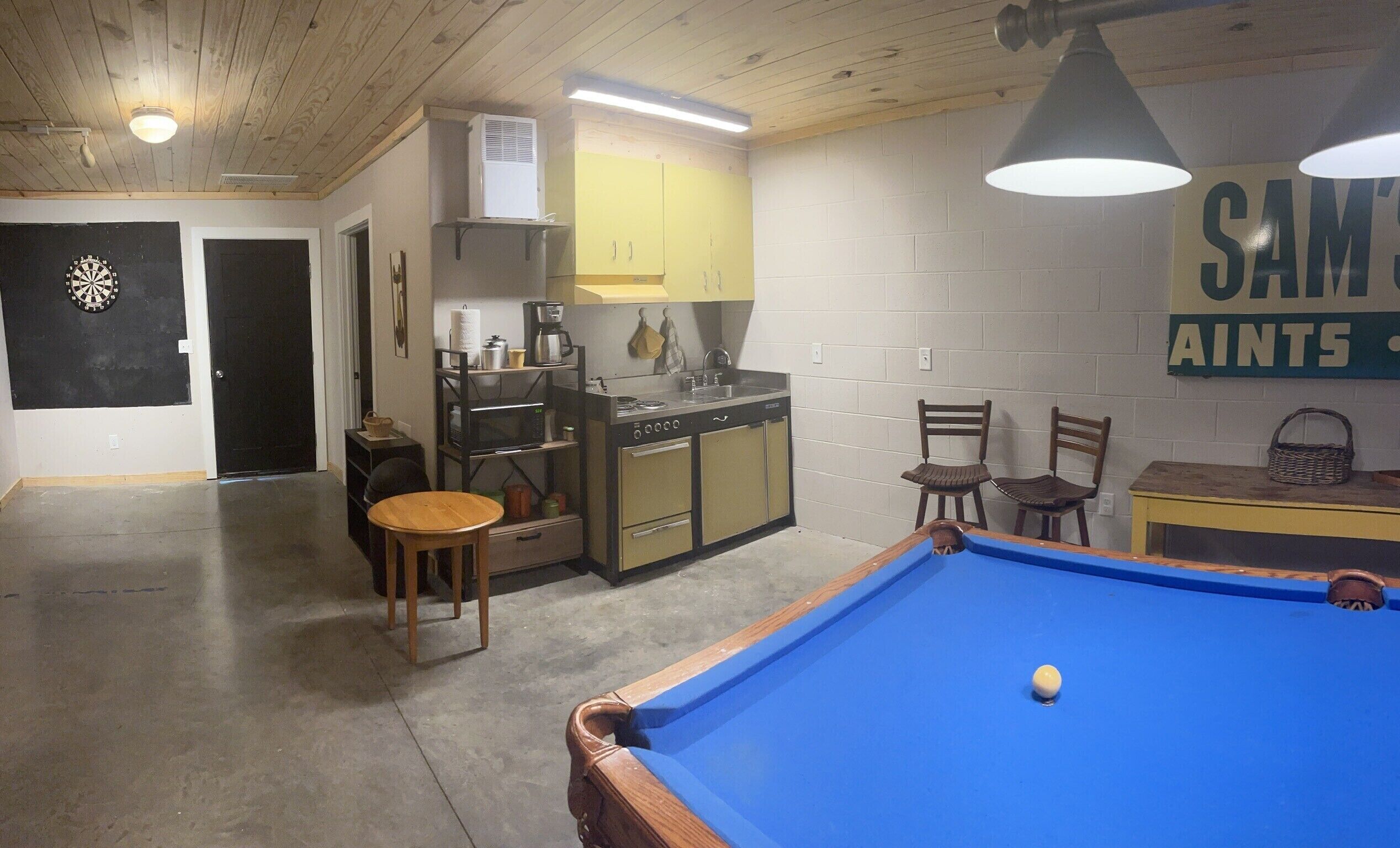 A room with a pool table, a dartboard, a kitchen area, and a sign reading "SAM'S AINTS".