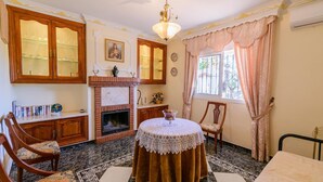 A room with a fireplace, wooden cabinets, a round table with a lace tablecloth, and a window with curtains.