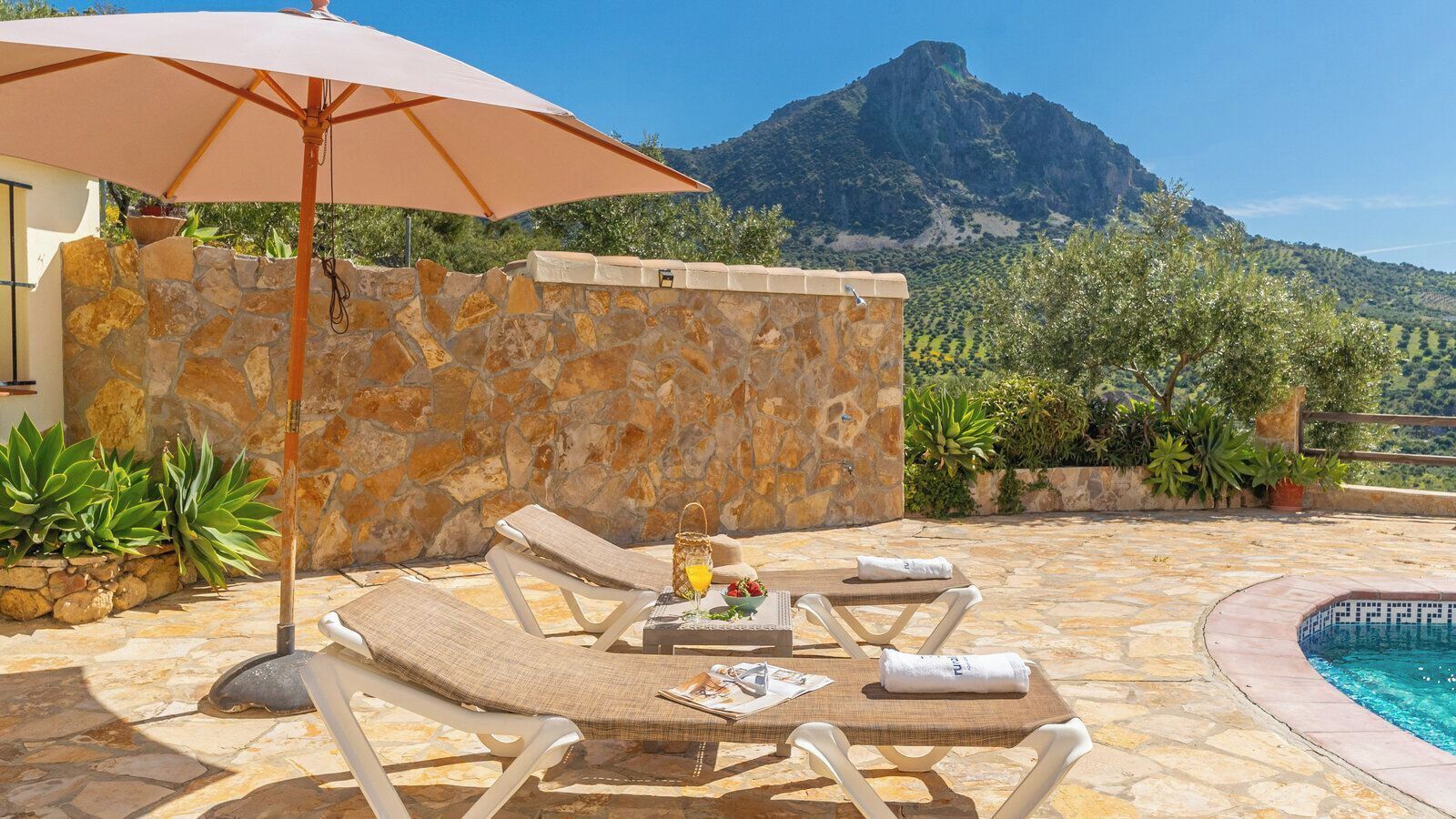 Zona de piscina con sillas de descanso, una mesa con una bebida y vistas a la montaña.