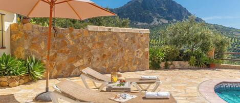 Zona de piscina con sillas de descanso, una mesa con una bebida y vistas a la montaña.