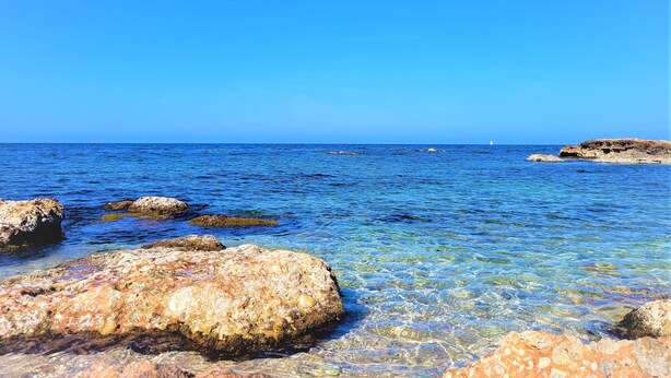Een heldere blauwe zee met rotsachtige oevers en een verre zeilboot.