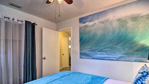 Un dormitorio con un cuadro grande de una ola en la pared, una cama con colcha azul y un ventilador de techo.