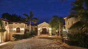 A well-lit, Spanish-style building with a cobblestone driveway, palm trees, and a stone pathway leading to the entrance. - Poolside Bliss Awaits, Cabo San Lucas Villa 1006 (Cabo San Lucas)