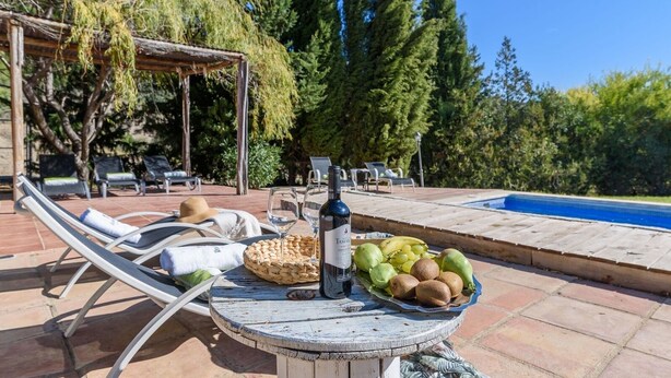 Un espace aménagé au bord de la piscine, avec des chaises longues, une table sur laquelle sont posés une bouteille et des fruits, et une pergola ornée de plantes grimpantes.