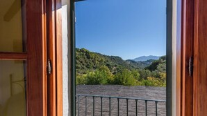 Une porte en bois ouverte laisse entrevoir un balcon d’où l’on peut admirer une colline boisée et des montagnes.