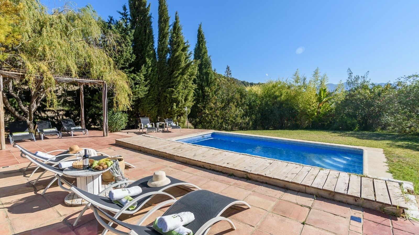 A swimming pool with lounge chairs and a pergola, surrounded by trees and a clear sky.