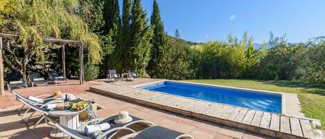 A swimming pool with lounge chairs and a pergola, surrounded by trees and a clear sky.