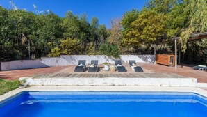 Une piscine avec quatre chaises longues, une terrasse en bois et un mur blanc en arrière-plan.