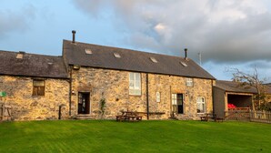A stone building with a sloped roof, multiple windows, and a grassy yard. - Bryn Lon Mawr: 5-Bed Barn Conversion in Caergeiliog | Managed by Anglesey Holiday Lettings (Caergeiliog)