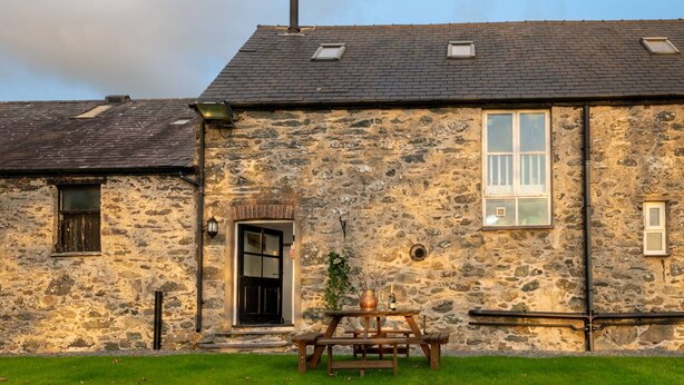 A stone building with a wooden door, a small window, and a picnic table with a vase and bottles. - Bryn Lon Mawr: 5-Bed Barn Conversion in Caergeiliog | Managed by Anglesey Holiday Lettings (Caergeiliog)