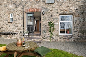 A stone building with a wooden table and chairs outside, a bottle of champagne, and a vase with branches.