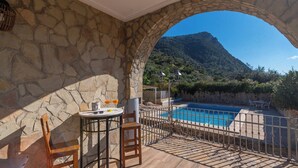 Un balcón de pared de piedra con piscina, sillas de madera y una mesa con bebidas.