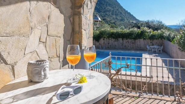 Una mesa de mármol blanco al aire libre, con dos vasos de jugo de naranja, una escultura de cabeza de Buda y un par de gafas de sol, con vistas a una piscina y un paisaje montañoso.