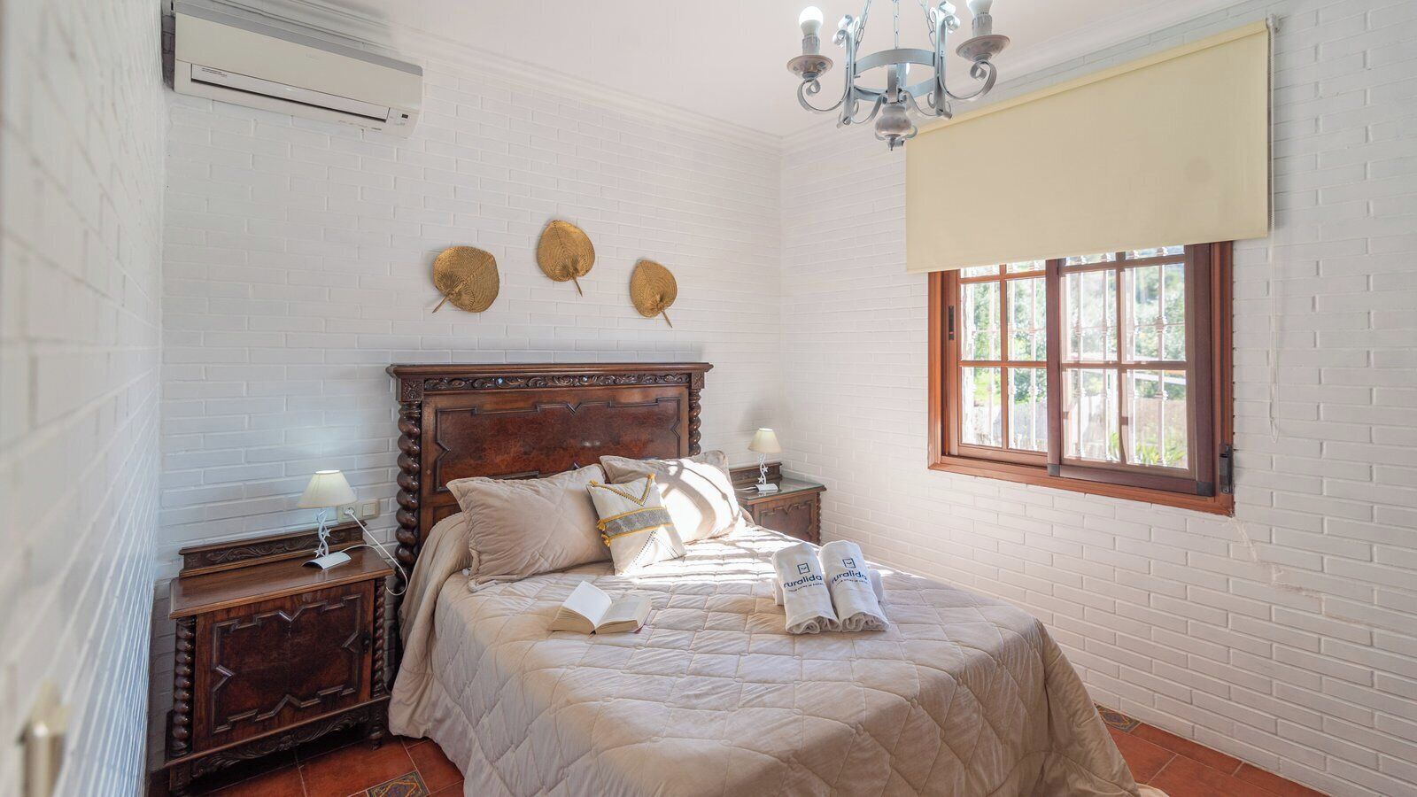 A bedroom with a wooden bed, a nightstand, a bedside lamp, a window with a view, and a chandelier.