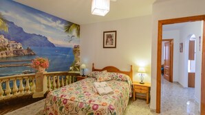 A bedroom with a bed, a nightstand, a lamp, a picture on the wall, and a view of the sea through a balcony.