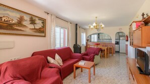 A living room with a red sofa, a small table, a TV, and a kitchen in the background.