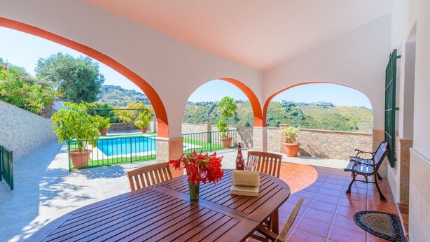 A patio with a wooden table, chairs, and a view of a swimming pool and greenery.