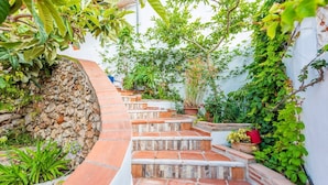 Degraus de pedra levam a uma parede branca, rodeada por vegetação exuberante e plantas em vasos.