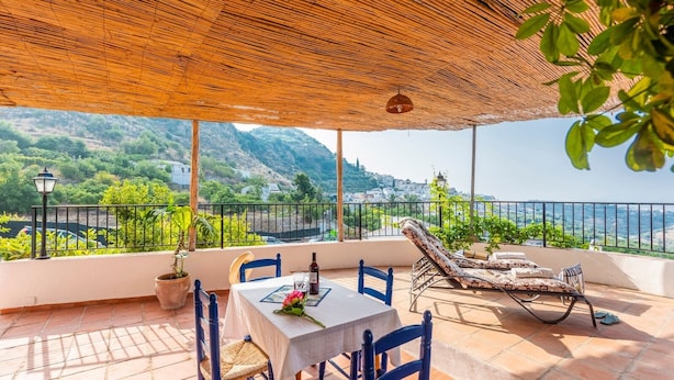 Terraço com mesa posta para dois, uma cadeira e espreguiçadeira, com vista para colinas e um corpo d'água.