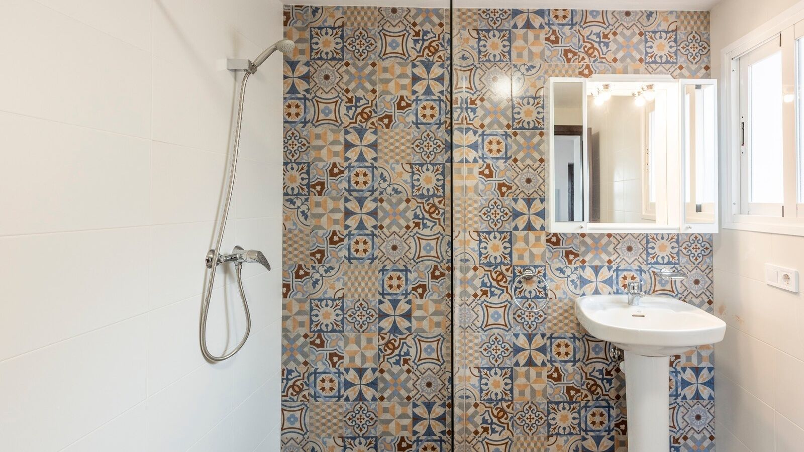 Een badkamer met een douche, een wastafel en een spiegel. De muren zijn betegeld met een decoratief patroon.