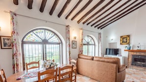 Uma sala de estar acolhedora com lareira, um sofá e uma mesa de jantar com cadeiras. Há grandes janelas com cortinas florais, e uma TV está fixada na parede.