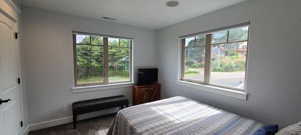 A bedroom with two windows, a bed, a bench, and a television. - Beautiful Home with Ocean View, Large Yard, and Hot Tub (Lincoln City)