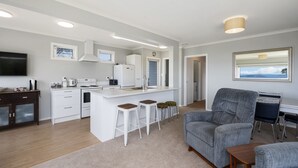 A modern kitchen with white cabinetry, a central island with stools, a dining area with a grey sofa, and a flat-screen TV. - I See The Lake - Omori Holiday Home (Omori)
