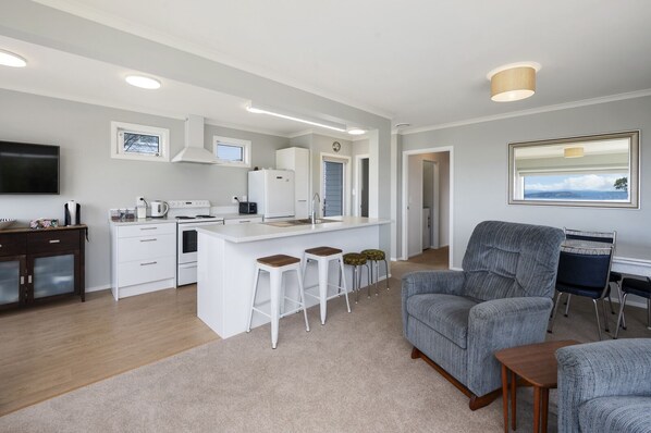 A modern kitchen with white cabinetry, a central island with stools, a dining area with a grey sofa, and a flat-screen TV.