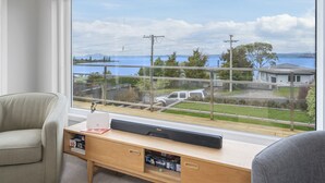 A room with a large window overlooking a body of water and a residential area. There is a desk with a laptop and a speaker, and a chair is visible to the left. - I See The Lake - Omori Holiday Home (Omori)